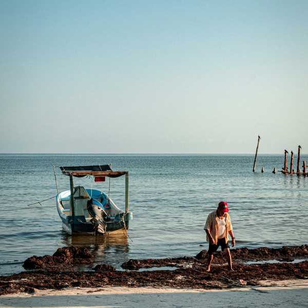 a man sitting on the shore of a beach next to a boat.