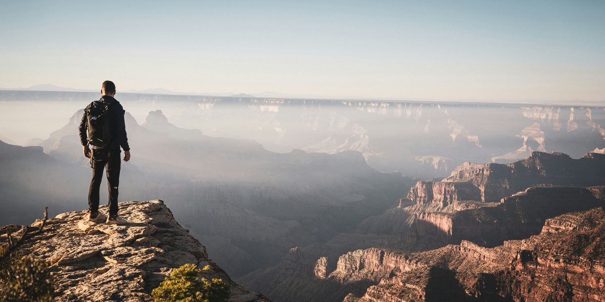A man stands on a rock overlooking a hazy Grand Canyon on a sunny day.