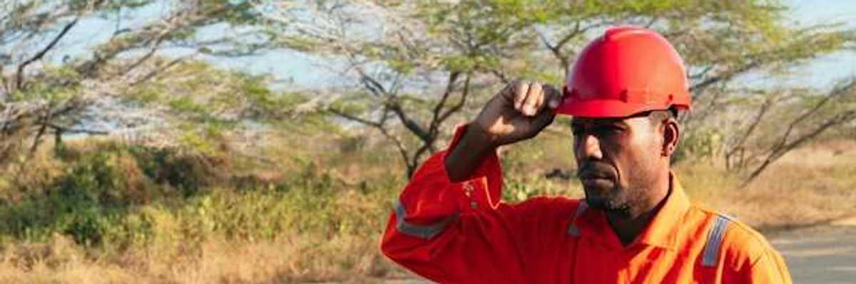 A man wearing an orange work jumpsuit and hard hat walks down a dirt road