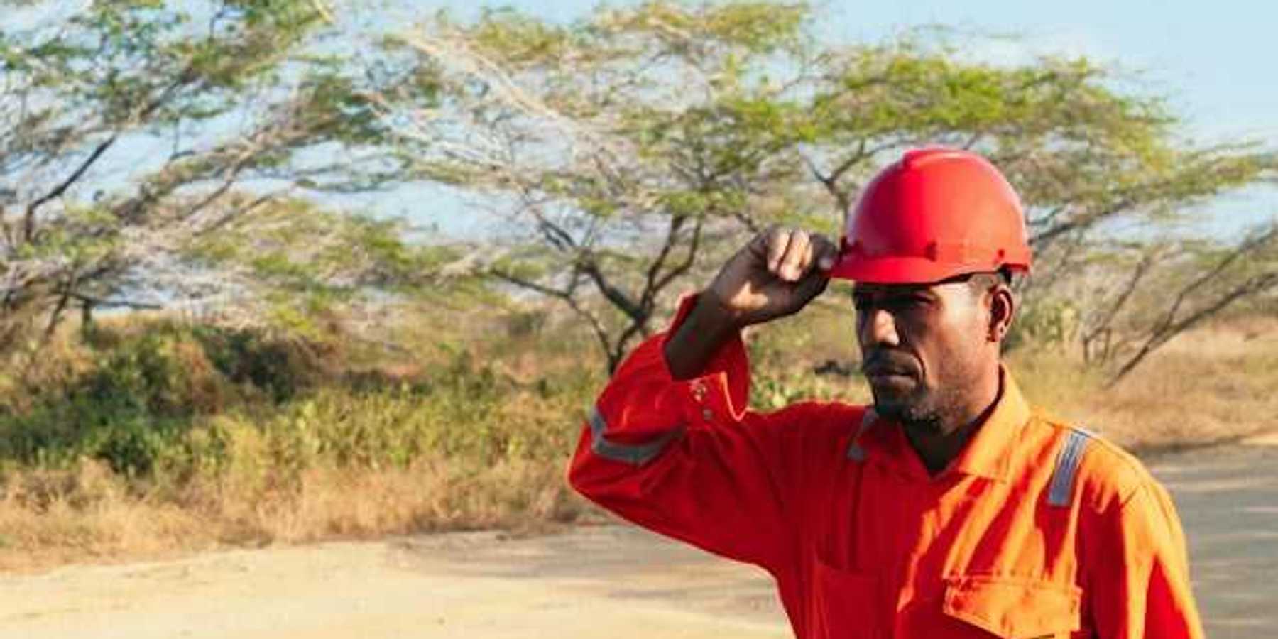 A man wearing an orange work jumpsuit and hard hat walks down a dirt road
