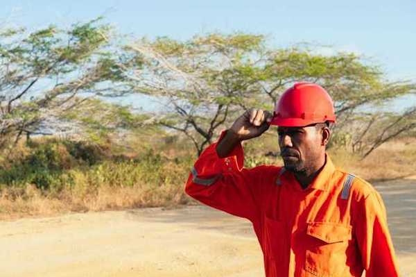 A man wearing an orange work jumpsuit and hard hat walks down a dirt road
