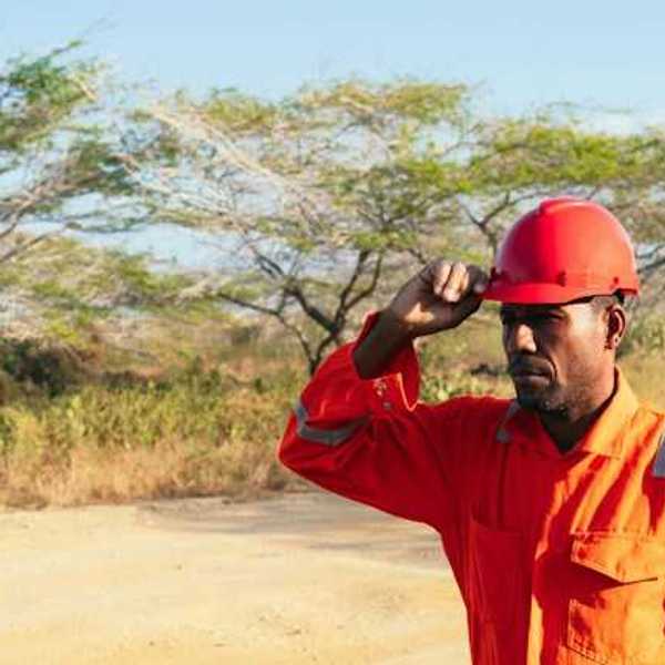 A man wearing an orange work jumpsuit and hard hat walks down a dirt road