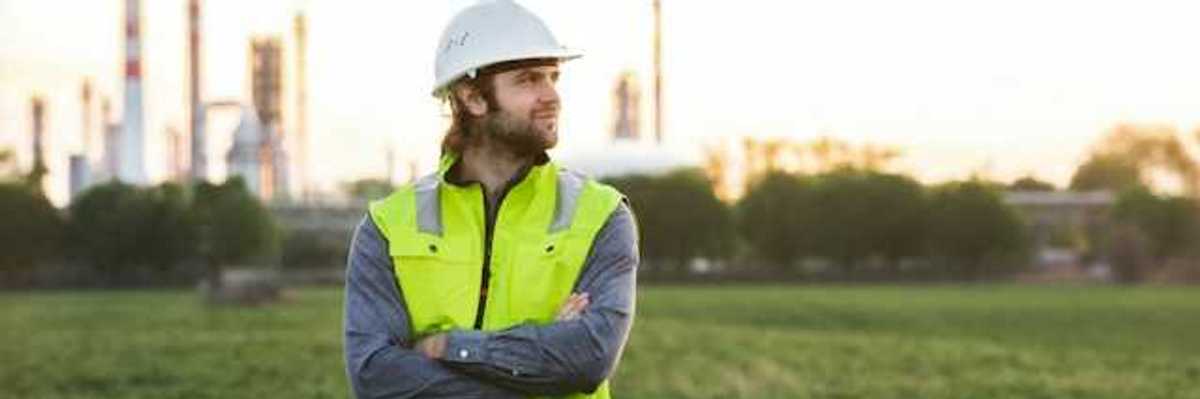 A man with a hard hat and neon vest on, posing in front of an oil refinery