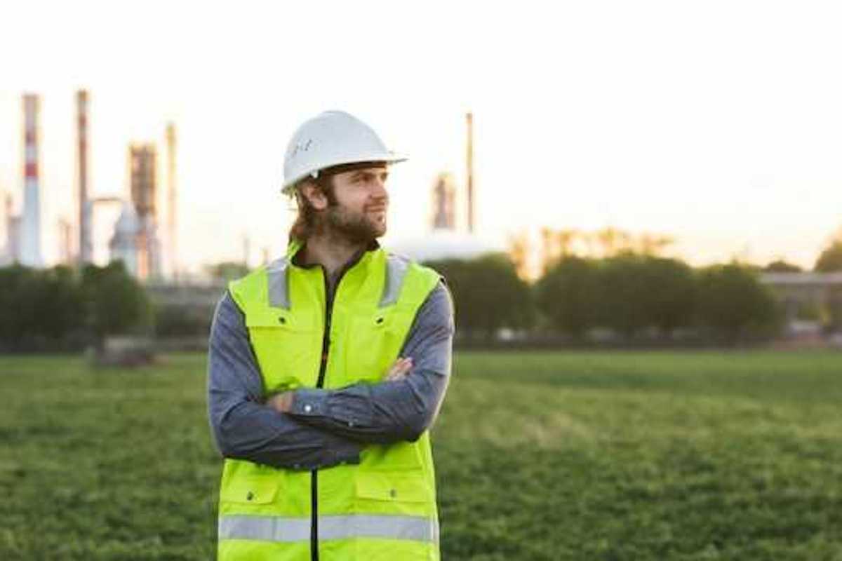 A man with a hard hat and neon vest on, posing in front of an oil refinery