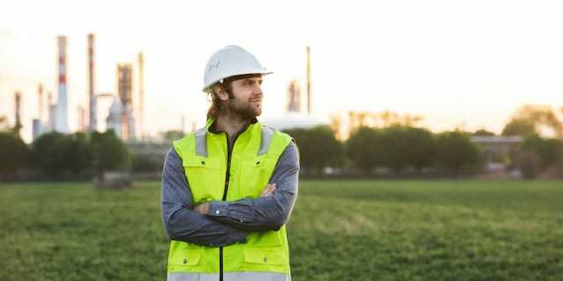 A man with a hard hat and neon vest on, posing in front of an oil refinery