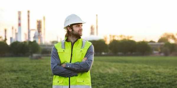 A man with a hard hat and neon vest on, posing in front of an oil refinery