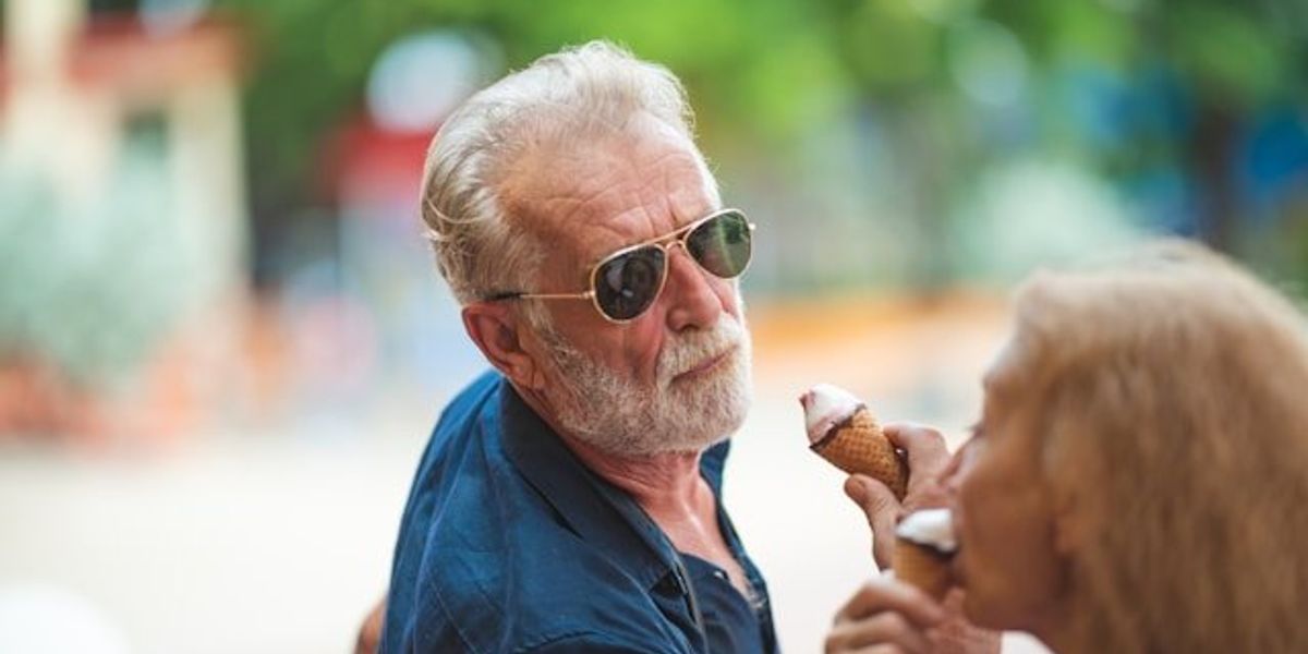 A middle aged man with a white beard eating an ice cream cone on a sunny day.