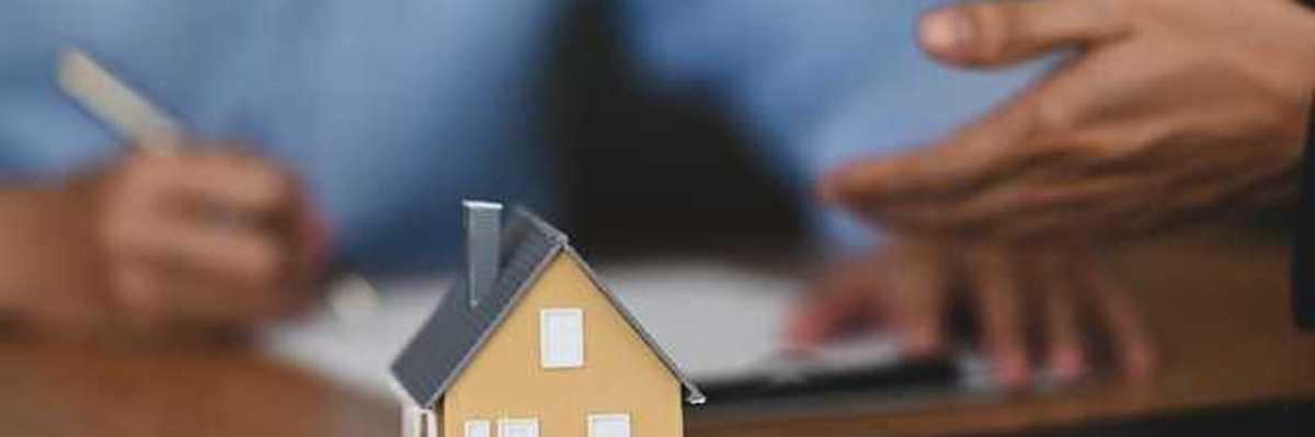 A model of a small house with two people signing paperwork in the background