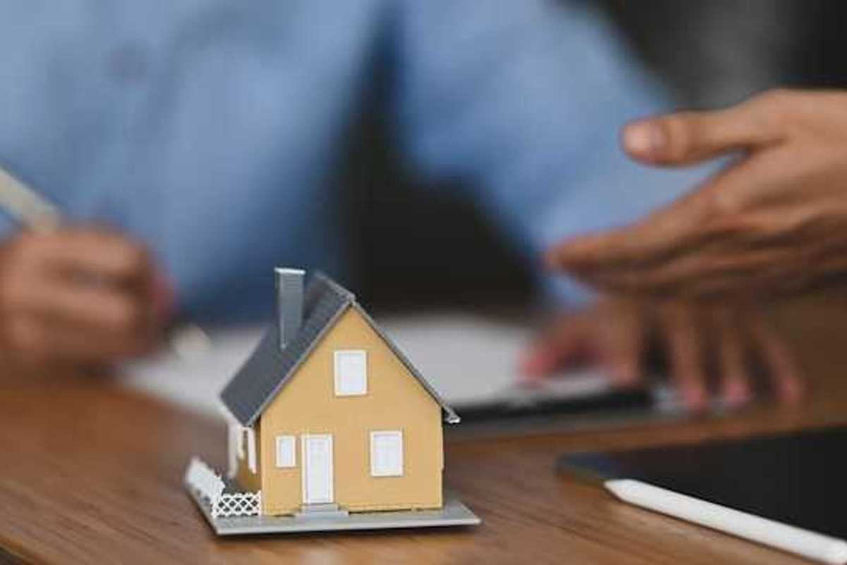 A model of a small house with two people signing paperwork in the background