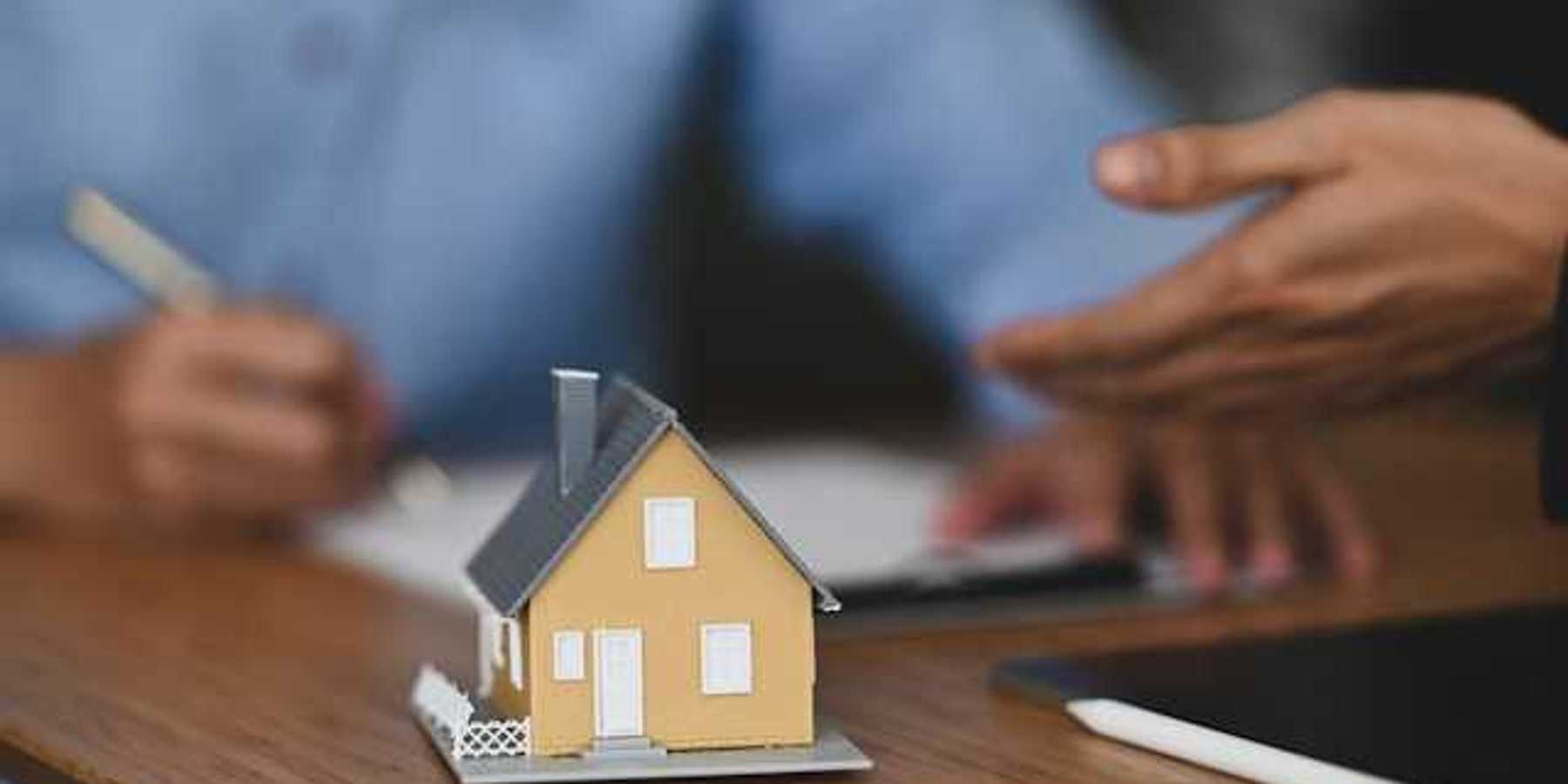 A model of a small house with two people signing paperwork in the background