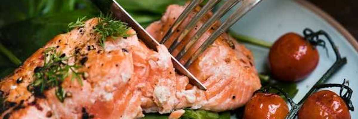 A person cutting a salmon filet on a plate with spinach and cherry tomatoes
