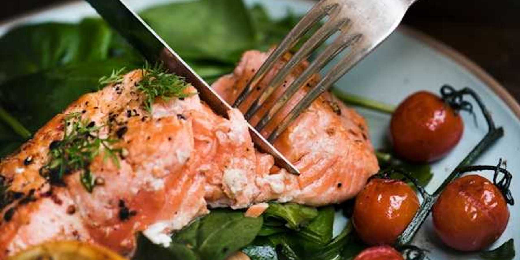 A person cutting a salmon filet on a plate with spinach and cherry tomatoes