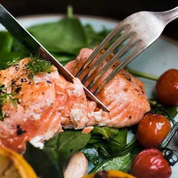 A person cutting a salmon filet on a plate with spinach and cherry tomatoes