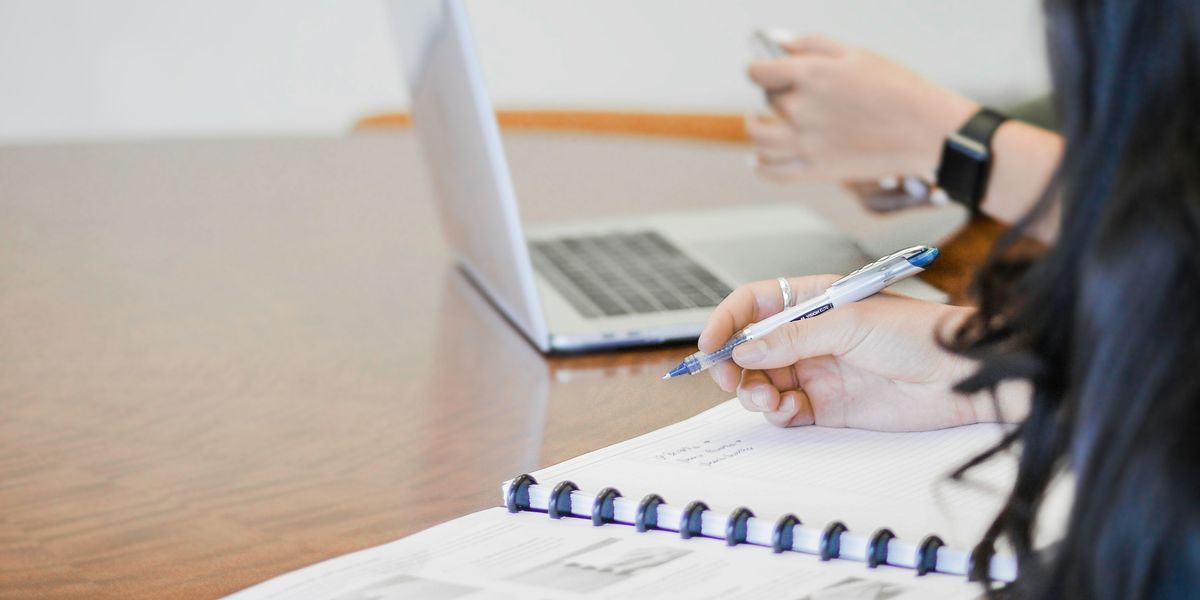 A person's hand holding pen writing on paper next to a person with a laptop.