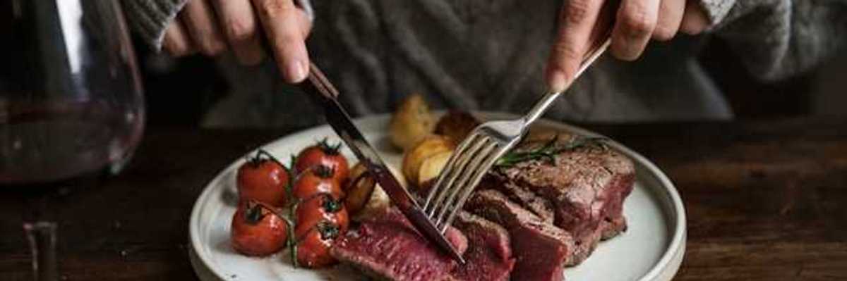 A person sitting at a table cutting into a cooked steak on a plate