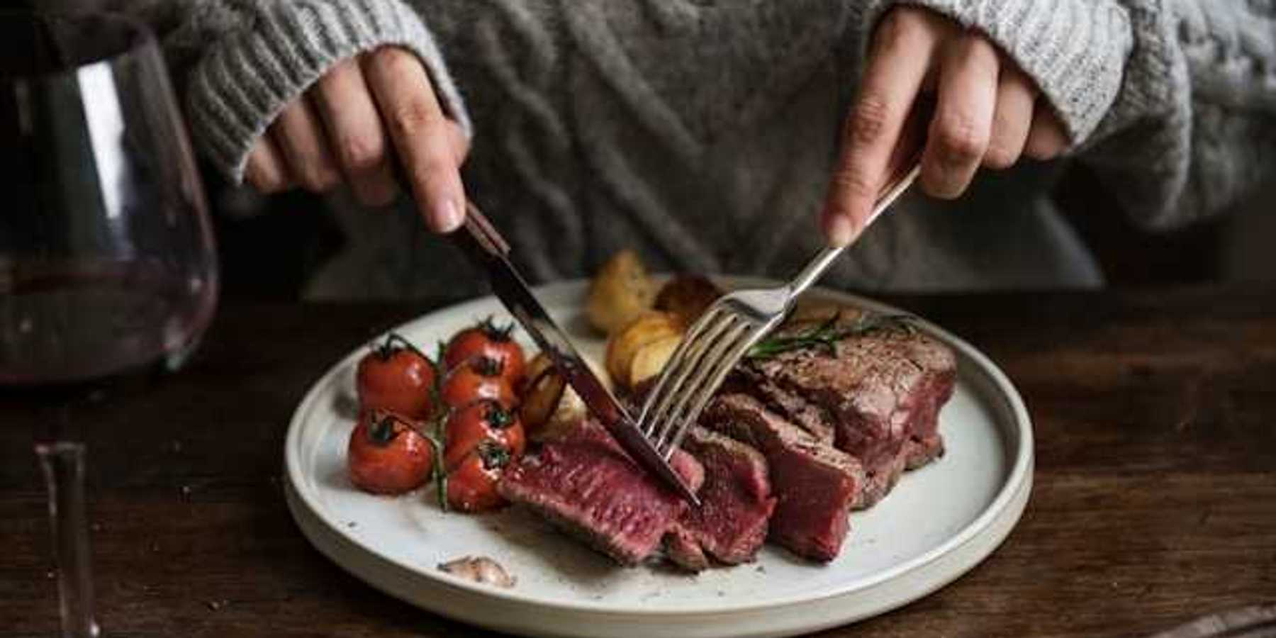 A person sitting at a table cutting into a cooked steak on a plate