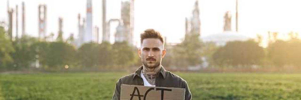 A person standing in front of a power plant holding a sign saying Act Now