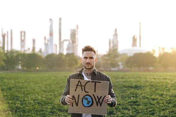 A person standing in front of a power plant holding a sign saying Act Now
