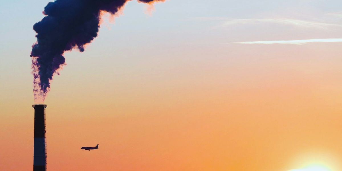 A power plant emitting pollution is silhouetted against a setting sun alongisde a plane and city skyline.