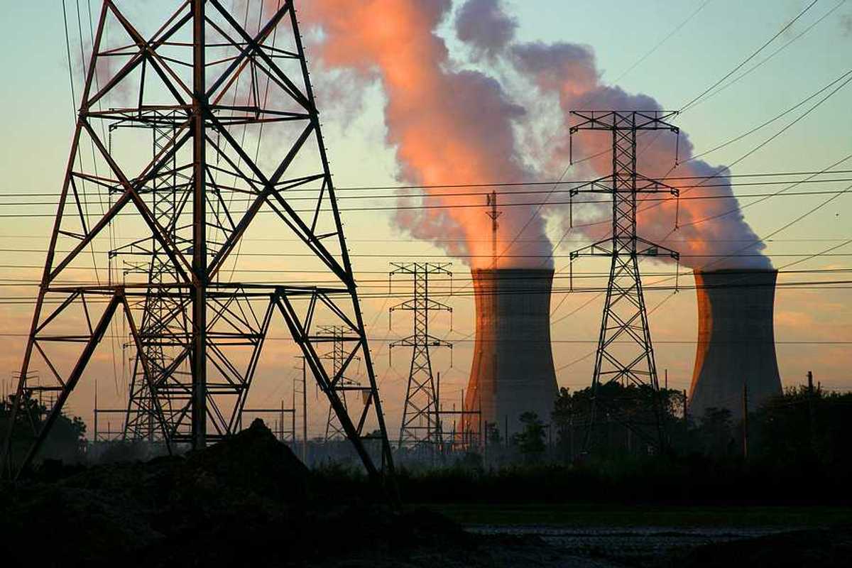 A power plant with smoke billowing from towers with electric energy lines in the foreground