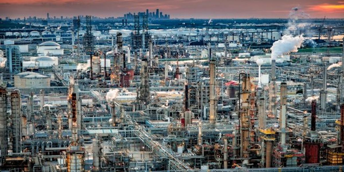 A refinery with smoke emitting from smokestacks with a city in the background.