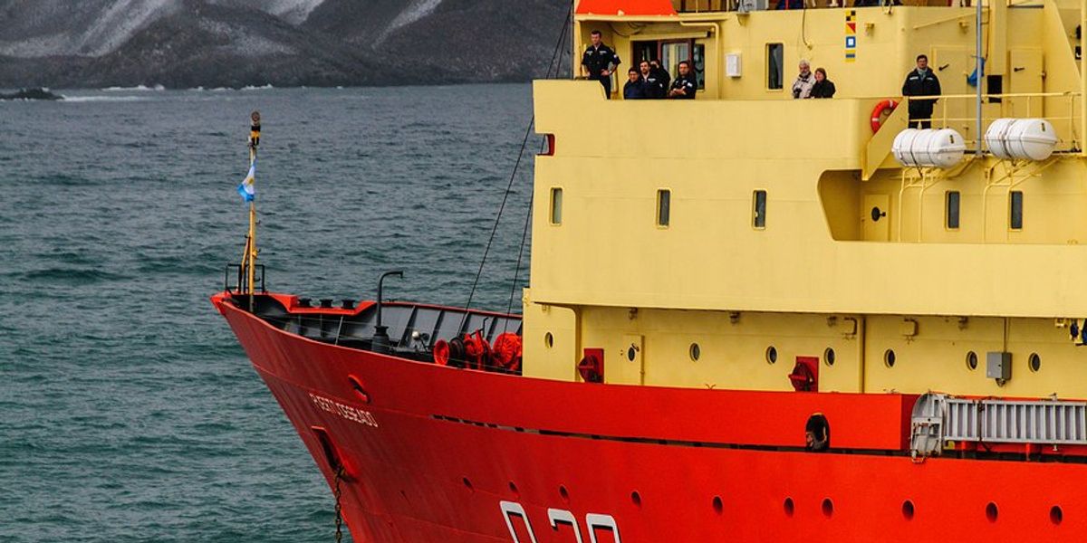 A research vessel in the sea with icy mountains in the background.