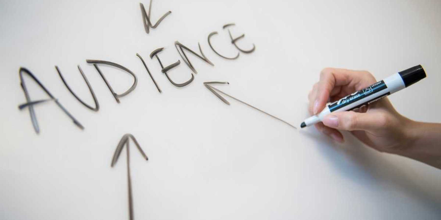 A right hand printing "AUDIENCE" with three arrows on a white board with a black marker