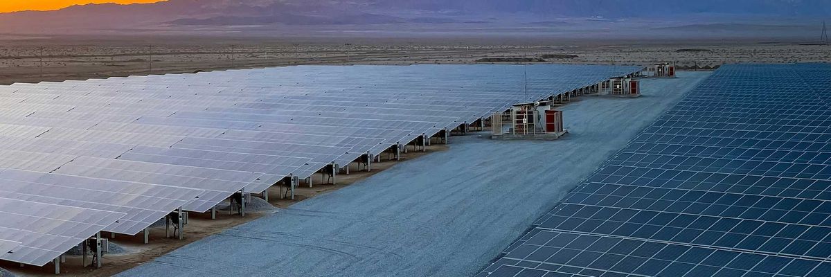a row of solar panels sitting on top of desert land with mountains and sunset in background.