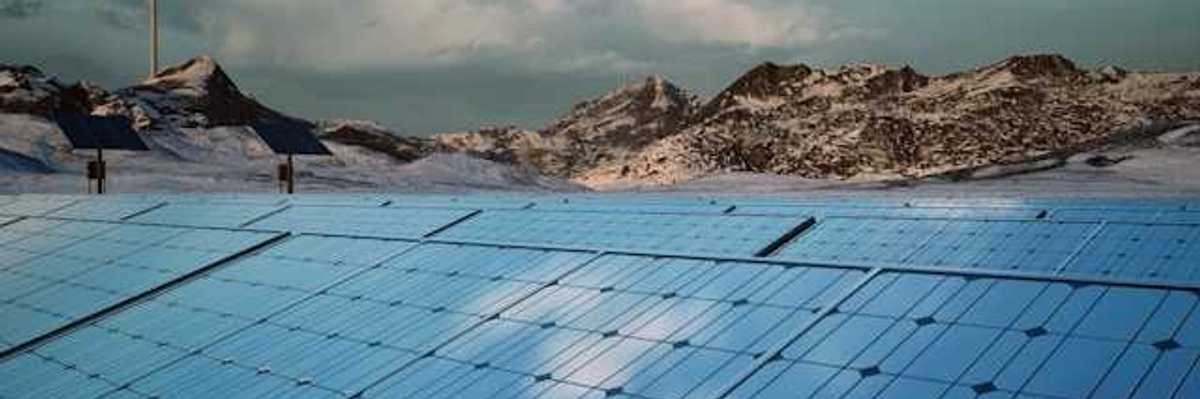 A row of solar panels with mountains and a wind turbine in the background