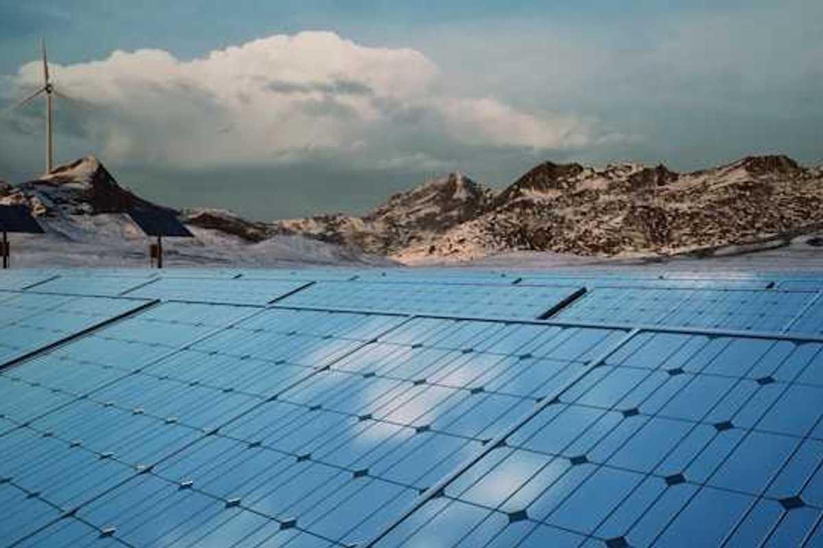 A row of solar panels with mountains and a wind turbine in the background