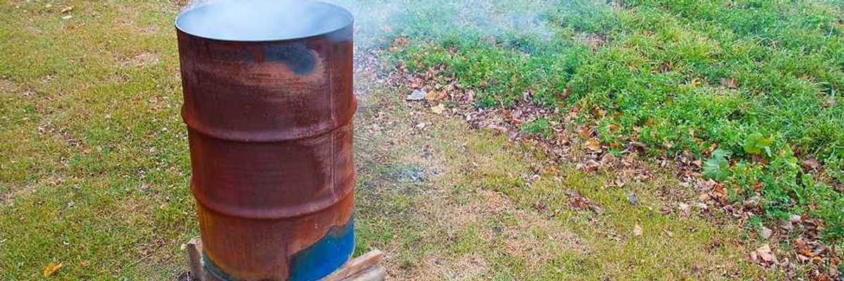 A rusty barrel with burning items in it, smoke billowing from the top