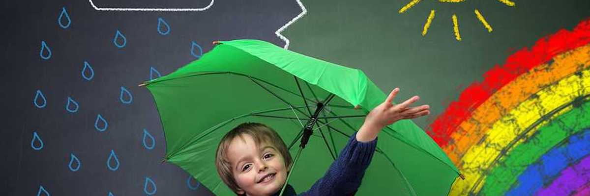 A small child under a green umbrella against a chalkboard with a rainbow and sun and rain on it