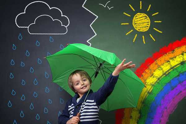 A small child under a green umbrella against a chalkboard with a rainbow and sun and rain on it