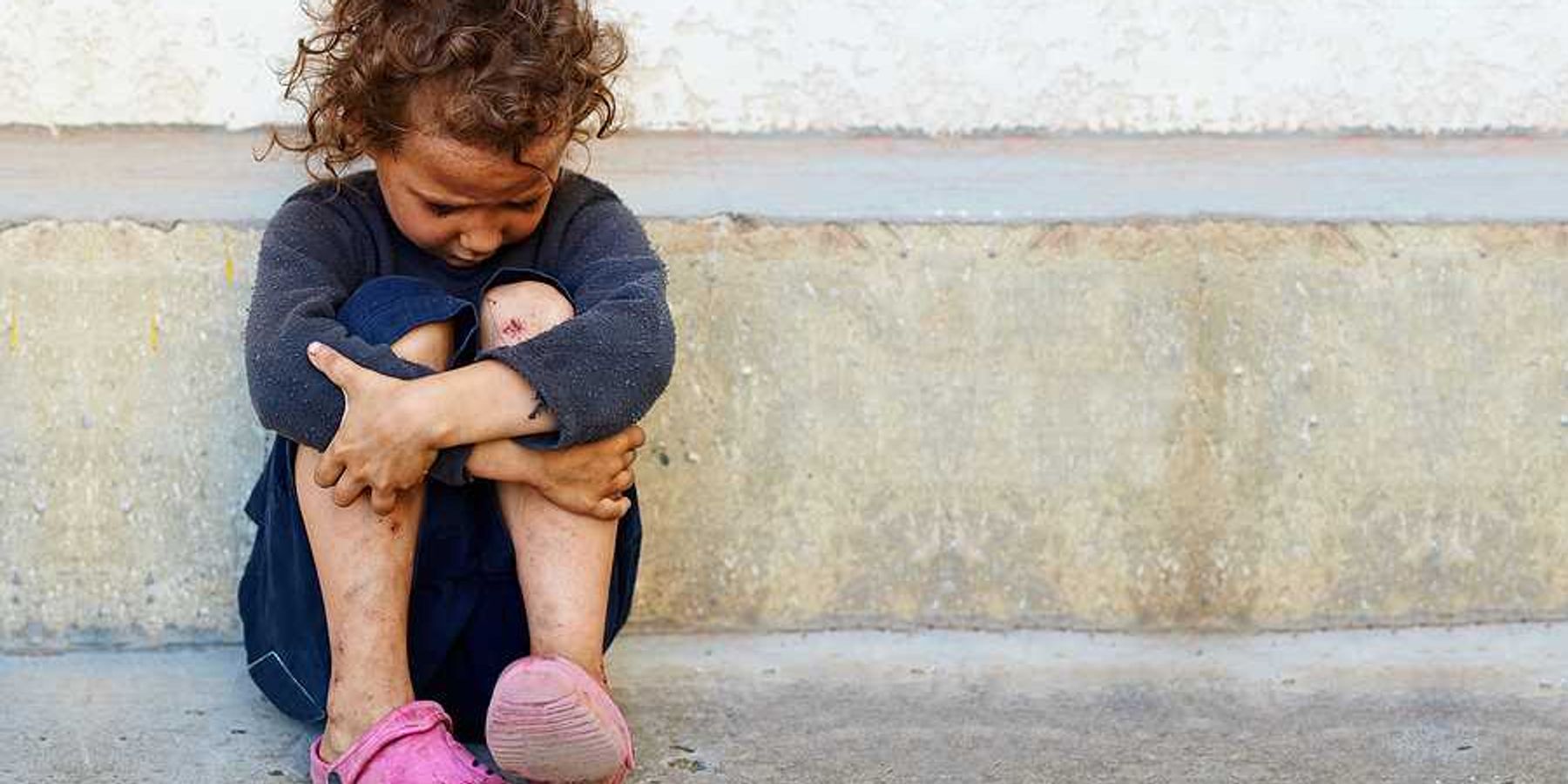 A small impoverished child sitting against a concrete curb