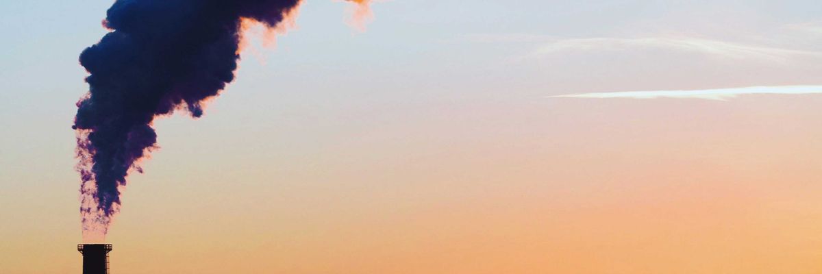 A smokestack spews pollution as a plane flies by in the distance across a cityscape and late day sun.