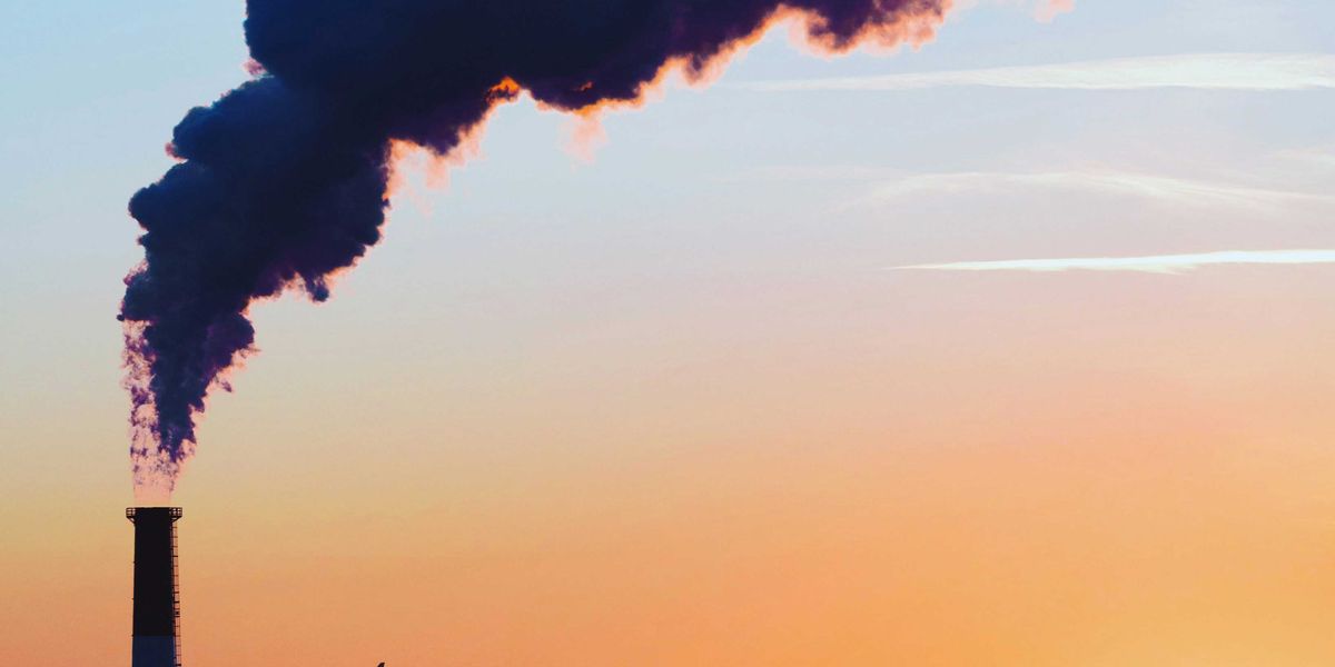 A smokestack spews pollution as a plane flies by in the distance across a cityscape and late day sun.