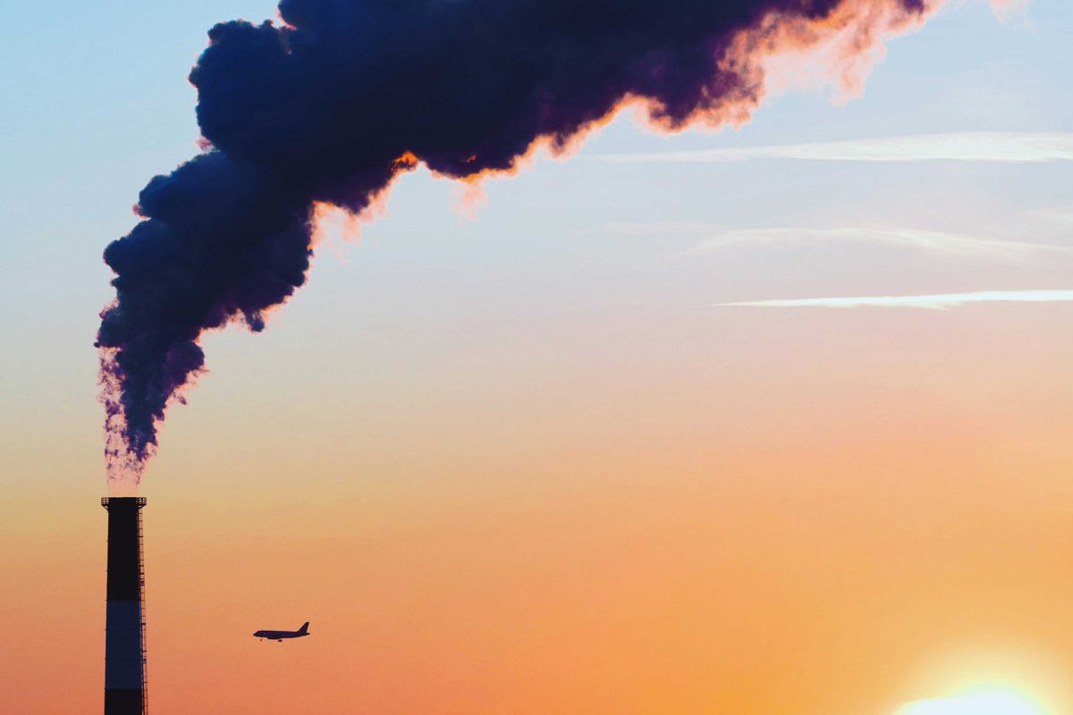 A smokestack spews pollution as a plane flies by in the distance across a cityscape and late day sun.