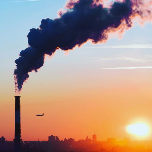 A smokestack spews pollution as a plane flies by in the distance across a cityscape and late day sun.