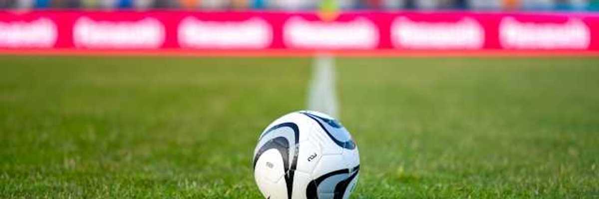 A soccer ball on green grass with people seated in bleachers in the background