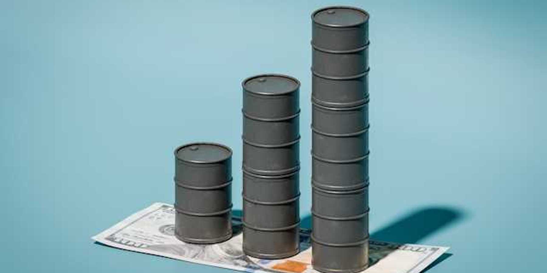 A stack of three oil barrels on top of a one hundred dollar bill against a blue background