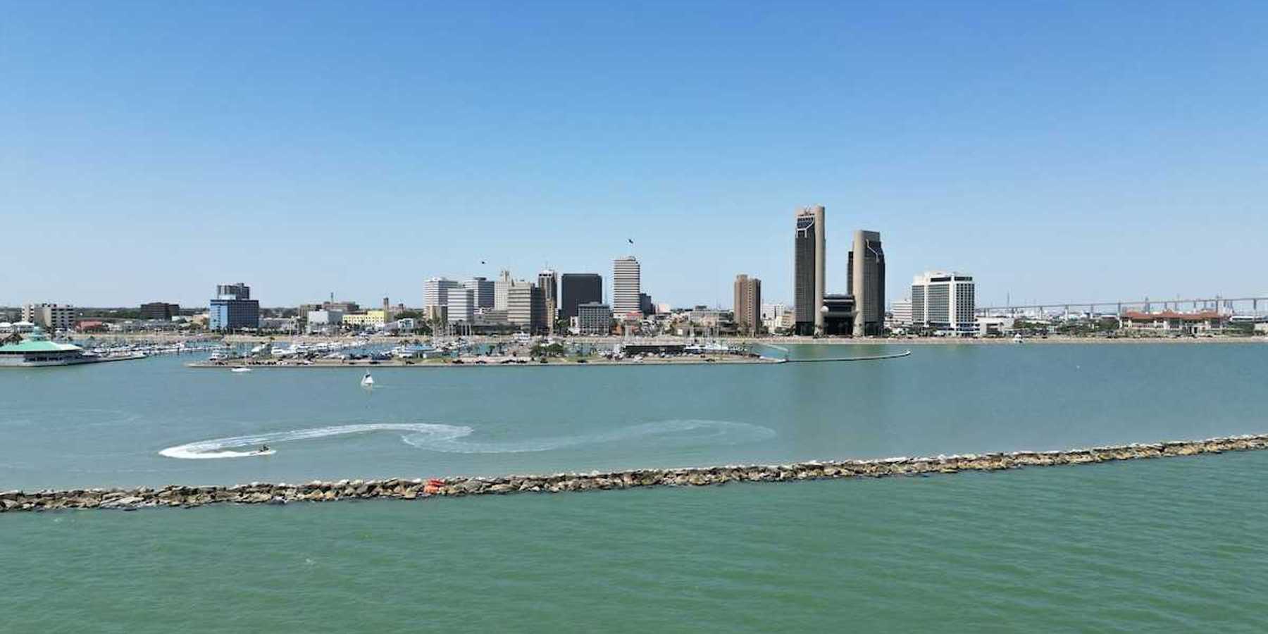 A sunny aerial view of downtown Corpus Christi, Texas, with the sparkling bay in the foreground and boats cruising through the water.