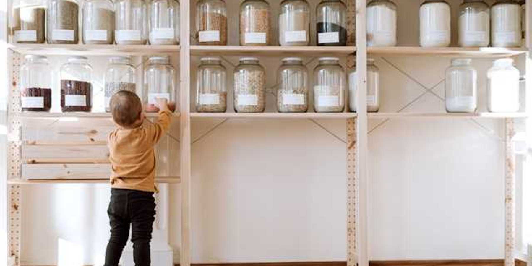 A toddler reaching up to a pantry shelf where jars of non perishable food is stored