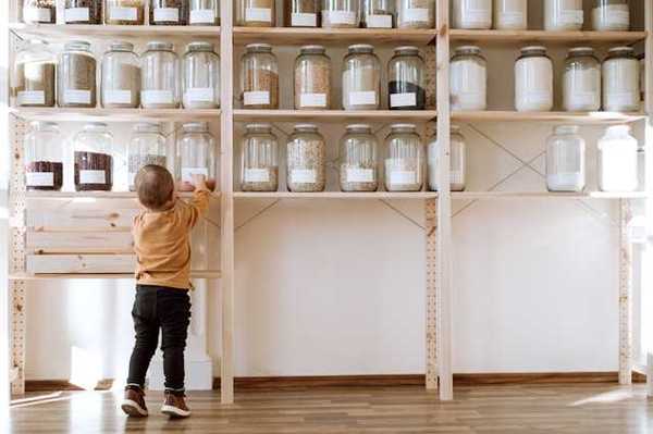 A toddler reaching up to a pantry shelf where jars of non perishable food is stored