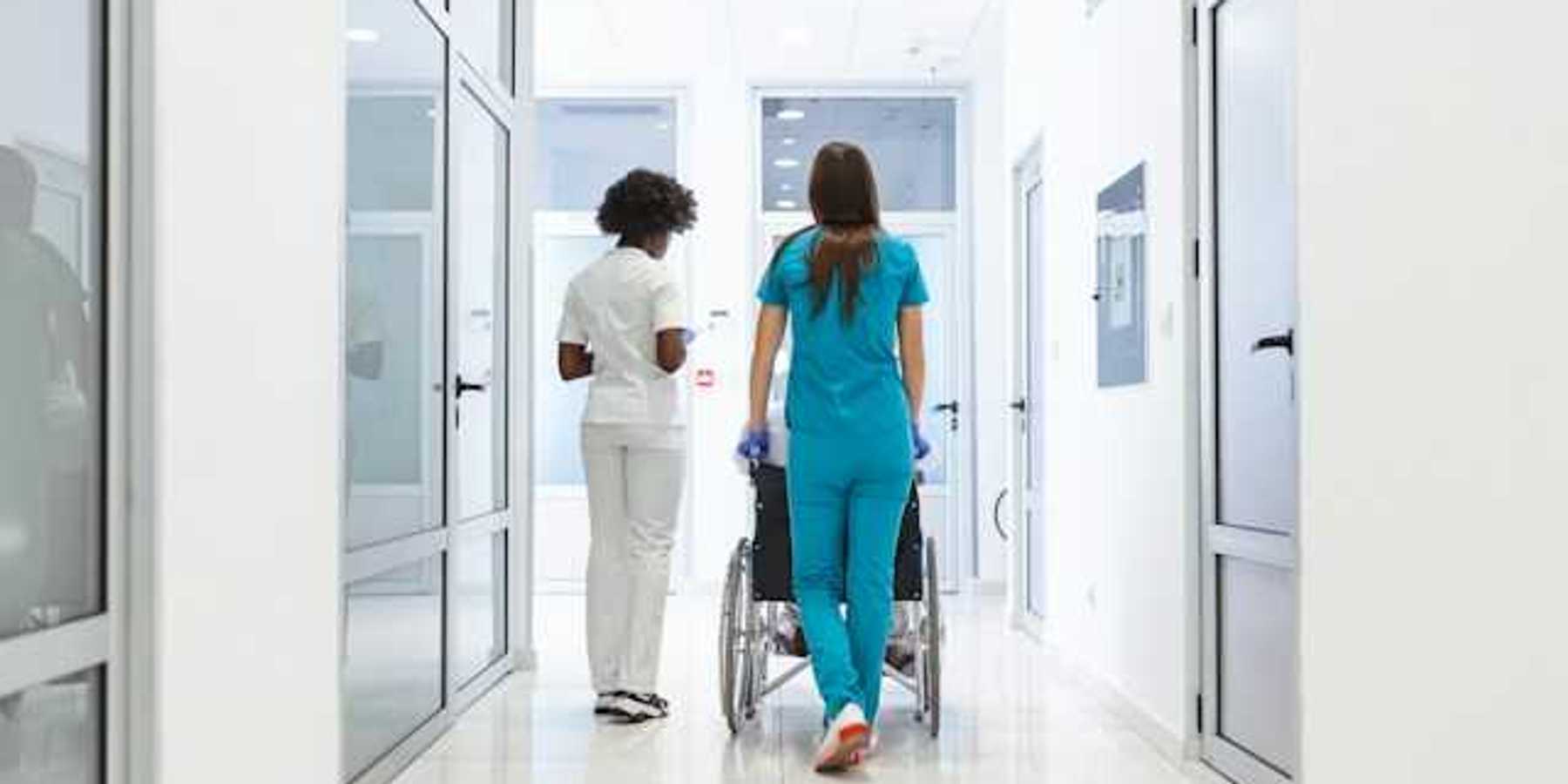 A view of a hospital corridor with two nurses wheeling a patient toward a room