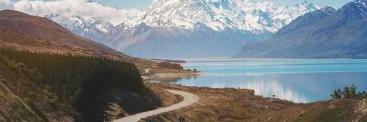 A view of a lonely highway next to an aqua lake with snow covered mountains in the background