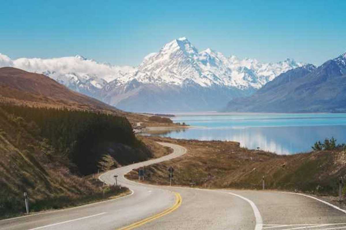 A view of a lonely highway next to an aqua lake with snow covered mountains in the background