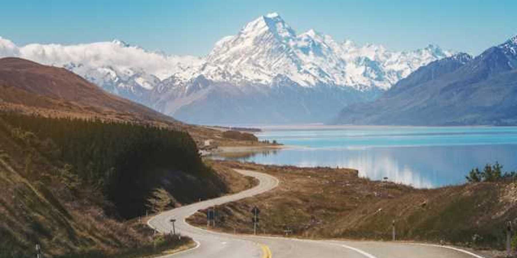 A view of a lonely highway next to an aqua lake with snow covered mountains in the background