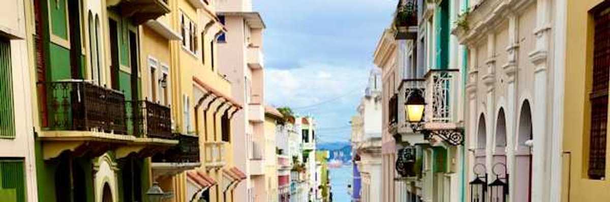 A view of a Puerto Rican street with multicolored buildings and cars and the ocean in the far distance
