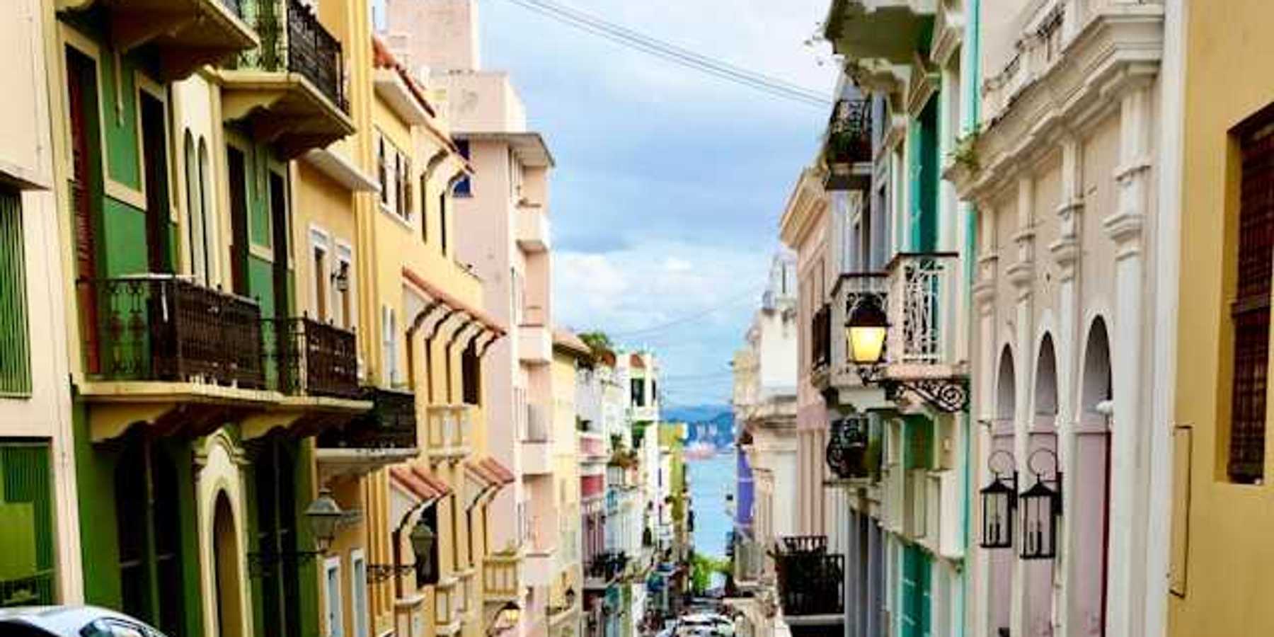 A view of a Puerto Rican street with multicolored buildings and cars and the ocean in the far distance