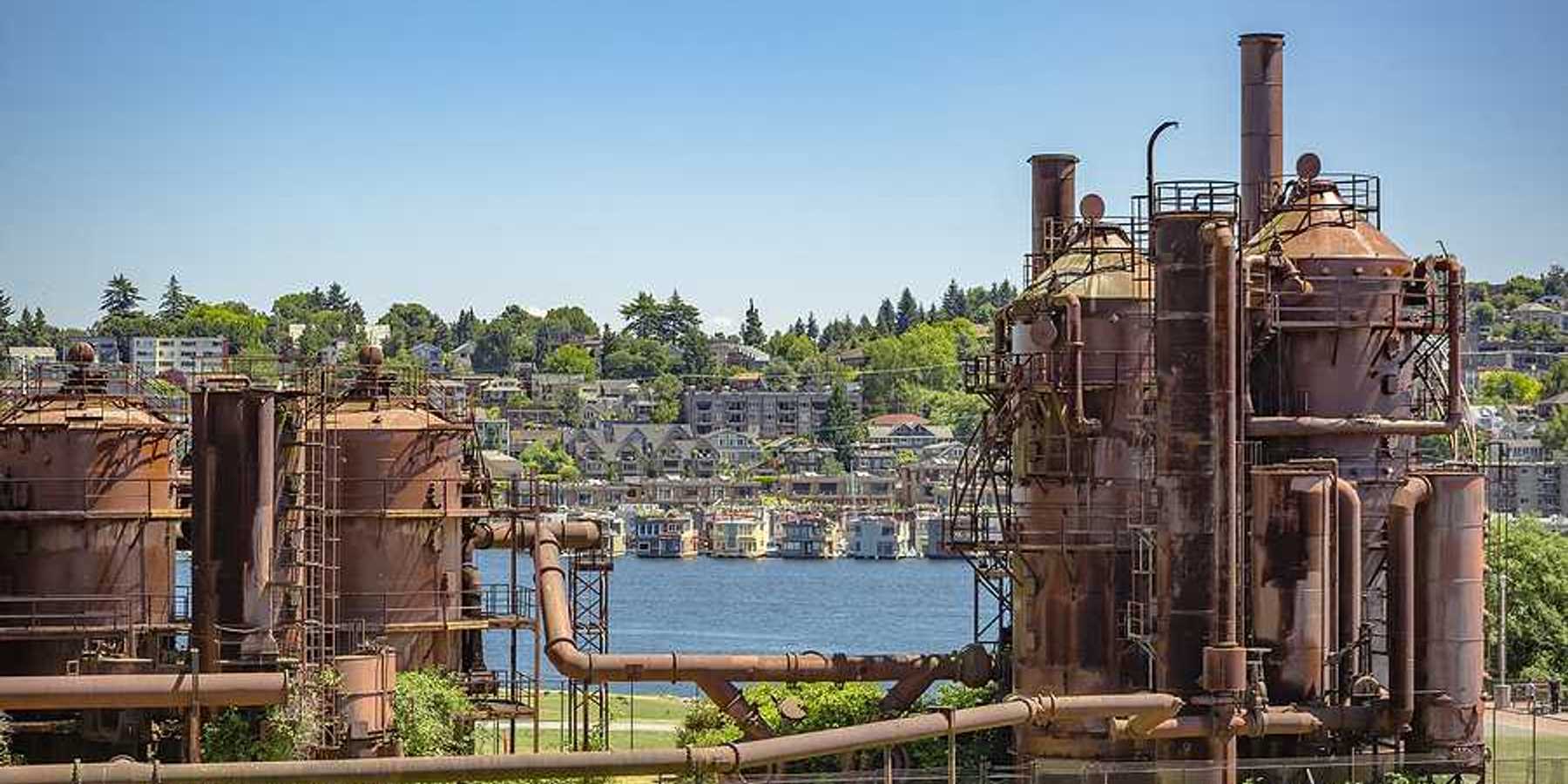 A view of rusting gas plant equipment on a green landscape with houses in the hills behind